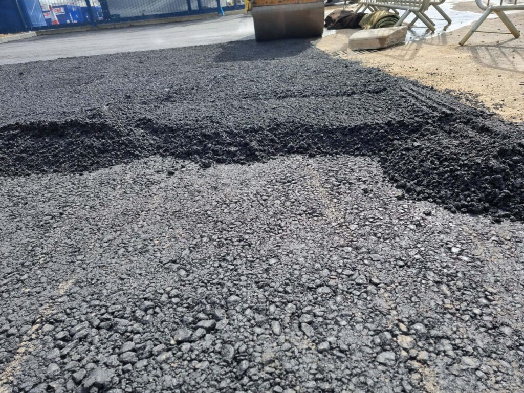 Dit is een foto van het aanleggen van een asfaltoppervlak. Op de achtergrond is een wals te zien.